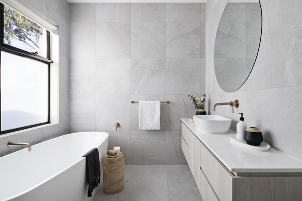 Badkamer met grote ronde spiegel en een witte waskom op een licht grijze wastafelmeubel met licht grijze marble tegels en een wit bad 